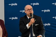 Mężczyzna w ciemnym garniturze i okularach - prezes Okręgowej Izby lekarskiej Artur Drobniak. W prawej dłoni trzyma mikrofon i przemawia. Za nim niebieska ścianka z białymi napisami.