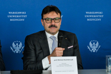 Mężczyzna w garniturze i krawacie siedzi przy stole konferencyjnym, trzymając mikrofon w ręce. 