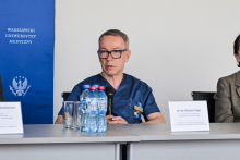 Mężczyzna w stroju medycznym siedzi przy stole. Przed nim stoją butelki z wodą i szklanki. Po lewej stronie widoczny niebieski baner z napisem „Warszawski Uniwersytet Medyczny” i emblematem. Na stole dwie tabliczki z nazwiskami, jedna z napisem „dr hab. Wojciech Figiel”.