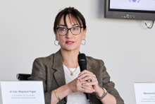 Kobieta w marynarce i białej bluzce siedzi za stołem, trzymając mikrofon w ręce.