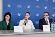 Trzy osoby siedzą przy stole z tabliczkami z imionami „Wiktoria” i „Danił”. Osoba pośrodku trzyma mikrofon. Na stole stoją butelki z wodą. W tle wielokrotnie powtórzone logo i napis „Warszawski Uniwersytet Medyczny”.