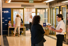 Dwie kobiety rozmawiające na korytarzu uczelni. Korytarz jest jasny i oświetlony. W tle widać inne osoby, część z nich siedzi przy stoliku. Obok nich niebieska ścianka z białymi napisami i logo Okręgowej Izby Lekarskiej.