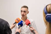 Młody człowiek w białej koszuli udziela wywiadu. Mówi do trzech mikrofonów różnych stacji radiowych i telewizyjnych.