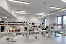 Kilka osób w białych fartuchach pracuje przy długim stole. Na blacie znajdują się butelki z odczynnikami, probówki, pipety i inne narzędzia. Uczestnicy mieszają substancje i zapisują wyniki. 