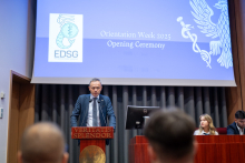Mężczyzna przemawia przy drewnianej mównicy. W tle ekran z napisem „Orientation Week 2025 Opening Ceremony” 
