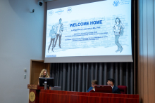 Kobieta stoi przy drewnianej mównicy. Na ścianie za nią wyświetlony jest slajd zatytułowany „Welcome home”.