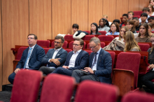 Grupa kobiet i mężczyzn siedzi w audytorium. W pierwszym rzędzie czterech mężczyzn ubranych w garnitury i krawaty. 