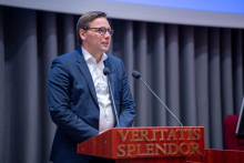 Mężczyzna w okularach, ubrany w niebieską marynarkę i białą koszulę, stoi za drewnianą mównicą.