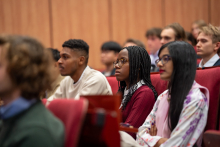 Grupa młodych ludzi siedzi w rzędach czerwonych foteli w sali wykładowej. Na pierwszym planie kobieta w bordowym swetrze z długimi splecionymi włosami i okularami w bordowym swetrze,