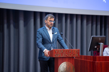 Mężczyzna w okularach, ubrany w elegancką marynarkę i koszulę, stoi za mównicą.