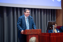 Mężczyzna w niebieskiej marynarce, białej koszuli i krawacie z wzorem przemawia przy drewnianej mównicy z napisem