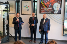 Trzech mężczyzn w garniturach: rzecznik WUM, rektor WUM i były rektor, stoi w jasnym pomieszczeniu. Osoba po lewej trzyma mikrofon i czyta z kartki. W tle widoczny ekran z portretem mężczyzny i napisem „Andrzej Kraśnicki 1949–2025” oraz ramki ze zdjęciami i tekstem.