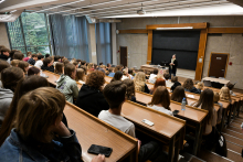 Grupa osób siedzących w drewnianych ławkach w auli wykładowej. Na środku auli stoi kobieta, prodziekan wydziału, która przemawia.