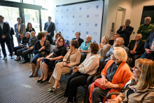 Grupa elegancko ubranych. Część z nich siedzi, część stoi. Znajdują się w jasnym pomieszczeniu z dużymi oknami. W tle widoczna ścianka z logotypami WUM. Osoby siedzące zwrócone są w stronę kobiety, która gestykuluje ręką. Wszyscy się uśmiechają.