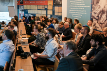 Grupa osób, które siedzą w sali. Osoby są ubrane formalnie i nieformalnie. Patrzą przed siebie, wyglądają, jakby czegoś słuchały. Niektóre osoby coś notują. Ściany w sali jest wyklejona kolorowymi tablicami ingformacyjnymi.