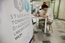 Młody mężczyzna siedzi przy stole i patrzy przez mikroskop. Obok, po lewej stronie, stoi rollup z napisem Studenckie Towarzystwo Diagnostów Laboratoryjnych WUM.