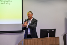 Elegancko ubrany mężczyzna, ubrany formalnie. Stoi przy białej ścianie z mikrofonem w dłoni. Przemawia. Obok niego widać fragment ekranu multimedialnego z wyświetlającą się prezentacją.