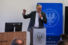 Mężczyzna w średnim wieku, formalnie ubrany, stoi przed mównicą. W lewej ręce trzyma mikrofon, prawą ma uniesioną do góry. Za nim stoi niebieski baner z logo WUM. Obok widać biurko z komputerem.