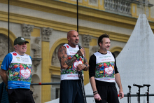 Trzech mężczyzn stoi na scenie przed budynkiem z żółtymi i białymi elementami. Noszą białe koszulki z kolorowym nadrukiem i napisem „Bieg po nowe życie”.