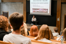 Grupa osób siedzących w auli. Patrzą przed siebie, w kierunku kobiety stojącej na środku auli. Kobieta jest elegancko ubrana, gestykuluje. Za nią widać ekran multimedialny z prezentacją o DNA.