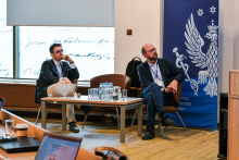 Dwóch formalnie ubranych mężczyzn. Siedzą na krzesłach na środku sali, między nimi widać stół. Głowy mają zwrócone w prawo, wyglądają jakby czegoś słuchali. Za nimi widać granatowy baner WUM z białym logo.