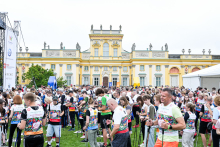 Około czterdziestu osób stoi na trawie przed żółto-białym budynkiem pałacowym z rzeźbami na dachu. Osoby są ubrane na sportowo i trzymają kijki do nordic walking.