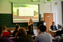 Grupa osób. Część z nich siedzi na krzesłach i patrzy przed siebie na stojących na środku: młodą kobietę i młodego mężczyznę. Za nimi widać ekran multimedialny z prezentacją. Obok przy komputerze siedzi jeszcze jedna młoda kobieta.