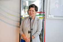 Kobieta, Karolina Skinder, ubrana elegancko stoi przed mikrofonem. Ma uśmiechniętą twarz. W tle widoczne są kolorowe wstążki: żółta, czerwona i zielona oraz fragment oprawionego zdjęcia na ścianie.