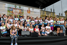 Około trzydziestu osób stoi na scenie pod zadaszeniem. Noszą kolorowe koszulki z logo „Bieg po Nowe Życie”. Uśmiechają się i machają do kamery. Za sceną znajduje się budynek z rzeźbami na dachu.