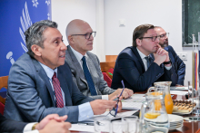 Trzy osoby siedzą przy stole w sali konferencyjnej. Mają na sobie eleganckie ubrania. Wyglądają, jakby słuchali w zamyśleniu. Na stole leżą dokumenty, długopisy, szklanki z wodą. W tle po lewej stronie widoczny jest niebieski baner z białymi symbolami i częściowo widocznym tekstem.