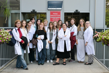 Grupa osób w białych fartuchach laboratoryjnych stoi przed wejściem do budynku oznaczonego tablicą z napisem „Wydział Farmaceutyczny”. Po obu stronach drzwi wejściowych ustawione są doniczki z kwiatami. 