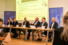 Grupa osób siedzących za stołem. Osoby są formalnie ubrane. Na stole widać białe kartki z wypisanymi nazwiskami i imionami tych osób. Jedna z osób, mężczyzna trzyma w ręce mikrofon i coś do niego mówi. Za nimi widać ekran multimedialny z wyświetlaną prezentacją. Są też niebieskie banery WUM z białym orłem.