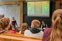 Grupa młodych osób siedzących w sali. Patrzą przed siebie, na dwie osoby stojące na środku. Po lewej stoi młoda kobieta, ubrana elegancko, po prawej elegancko ubrany mężczyzna. Mężczyzna trzyma w rękach tablet. Za nimi widać ekran z prezentacją "Apteka Wiedźmina". 