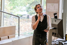 Młody mężczyzna, elegancko ubrany. Stoi, w prawej ręce trzyma mikrofon. Głowę ma uniesioną do góry i w prawo, spogląda na coś. Obok niego widać ekran komputera. Za nim wysokie okna.