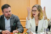 Dwie osoby siedzą przy stole i rozmawiają. Kobieta lewej ma szarą marynarkę i jasnoniebieską koszulę. Mężczyzna po prawej ma białą marynarkę i uniesione ręce. 