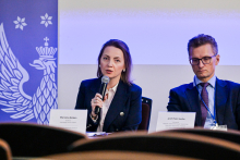 Dwie osoby siedzą przy stole podczas konferencji lub panelu. Osoba po lewej trzyma mikrofon. Przed nią leży tabliczka z napisem „Marzena Nielsen”. Po prawej stronie siedzi mężczyzna z tabliczką „prof. Piotr Socha”. Po lewej stronie zdjęcia widoczny jest niebieski baner z białym orłem i gwiazdkami.