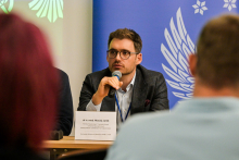 Mężczyzna siedzi przy stole i mówi do mikrofonu. Jest ubrany formalnie. Przed nim leży tabliczka z napisem „dr n. med. Maciej Janik” oraz informacją o jego afiliacji z European Reference Network Adult LCH, UZL. W tle widoczne są niebiesko-białe wzory z motywem skrzydeł i gwiazd. Na pierwszym planie znajdują się rozmyte sylwetki innych osób.