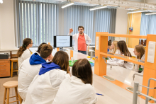 Grupa osób w laboratorium. Wszyscy są ubrani w białe fartuchy. Większość osób siedzi przy stołach. Jeden mężczyzna stoi, prawą rękę ma uniesioną do góry, przemawia.