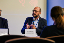 Mężczyzna w średnim wieku, ubrany formalnie, siedzi. W lewej ręce trzyma mikrofon, prawą gestykuluje. Obok widać fragment innej osoby, a po lewej fragment niebieskiego banera.