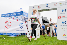 Kobieta w białej koszulce z kolorowym nadrukiem i czarnych legginsach przechodzi przez bramkę z logotypami sponsorów. Trzyma kijki do nordic walking. W tle widać namiot z napisem „Mamy w sobie niejedno życie” oraz baner z hasłem „Bieg po nowe życie”. 
