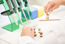 Na pierwszym planie widoczne są dłonie osoby manipulującej fiolką z żółtą cieczą. Na blacie znajdują się inne fiolki z substancjami, ołówek oraz pojemnik z napisem „chromatografia”. W tle można dostrzec stojak z pipetami oraz kolorowe pojemniki na próbki. Kompozycja obrazu skupia się na precyzyjnej pracy laboratoryjnej i ukazuje elementy typowe dla środowiska badawczego.