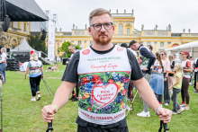 Mężczyzna w kolorowej koszulce z napisem „Bieg po nowe życie” i logo „Polska Transplantologia” stoi na trawie przed pałacem. Mężczyzna trzyma kijki do nordic walking. W tle widać innych uczestników wydarzenia, namioty i scenę. 