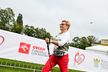Kobieta w średnim wieku idzie z kijkami do nordic walking. Ma krótkie blond włosy, okulary, białą bluzkę i czerwone spodnie. W tle widać baner z logotypem „ORLEN Fundacja im. Łukasiewicza” i napisem „Bieg po nowe życie”. Po lewej rosną drzewa, po prawej stoi namiot i budynek muzeum w Wilanowie. 