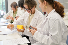 W jasnym pomieszczeniu laboratoryjnym cztery osoby w białych fartuchach siedzą przy stole, wykonując zadania praktyczne. Na pierwszym planie jedna osoba trzyma żółty pojemnik, a druga używa suwmiarki. Na stole leżą dokumenty i długopisy. W tle dwie kolejne osoby pracują nad swoimi zadaniami – jedna z nich trzyma urządzenie elektroniczne. 