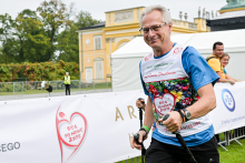 Mężczyzna w średnim wieku idzie z kijkami do nordic walking. Nosi niebieską koszulkę z kolorowym nadrukiem i napisem „Bieg po Nowe Życie”. W tle widać żółty budynek, biały namiot i baner z czerwonym symbolem serca oraz napisem „Bieg po Nowe Życie”.