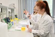 Grupa młodych osób w białych fartuchach laboratoryjnych. Siedzą przy stołach, w rękach trzymają pipety. Przed nimi widać materiały edukacyjne.