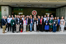 Około 30 osób stoi przed wejściem do budynku. Ubrani są formalnie lub półformalnie, część ma identyfikatory na smyczach. W tle widoczna jest ściana z dużym emblematem oraz dwa znaki po bokach. 