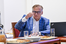 Mężczyzna siedzący przy stole. Jest elegancko ubrany i ma okulary. Gestykuluje jedną ręką, którą ma uniesioną w górę.