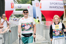 Mężczyzna w białej koszulce, zielonych spodenkach i czarnej czapce oraz kobieta w czarnej bluzie i białych spodniach idą w kierunku mety. Oboje noszą numery startowe z sercem i napisem „Bieg po Nowe Życie”. W tle widać balony z napisem „SPORTOWA WARSZAWA” oraz kibiców klaszczących za barierkami.