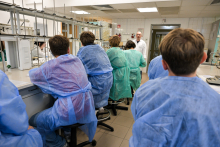 W laboratorium grupa osób siedzi na krzesłach laboratoryjnych, ubrana w niebieskie i zielone fartuchy ochronne. Są zwróceni plecami do obiektywu i słuchają osoby prowadzącej zajęcia, która stoi przed nimi w białym fartuchu. W tle widoczne są różne urządzenia i sprzęt laboratoryjny. 